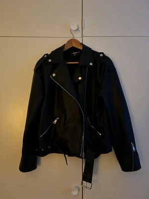 Svart  skinnjacka med bälte - Säljer en svart  skinnjacka  med silvriga dragkedjor, axelklaffar och bälte i midjan. Jackan har en klassisk oversized passform och långa ärmar. 