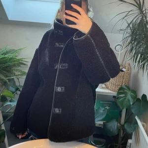 Svart teddyjacka med knappar - Mysig svart teddyjacka med stora knappar framtill och markerade sömmar i skinnimitation. Jackan har hög krage och lång ärm, den är i jättebra skick knappt använd! 