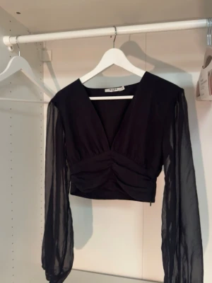Svart croppad topp från NA-KD - Snygg svart croppad topp från NA-KD med transparenta långärmade ärmar och v-ringad front. Även dragkejda på sidan. Toppen har en draperad detalj över bysten och är tillverkad i ett lätt, flowy material som ger en elegant look. Världens vackraste topp men tyvärr blivit för liten🥹 