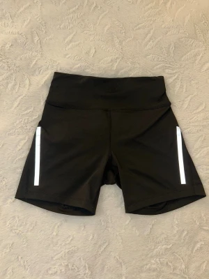 Svarta träningsshorts från SOC - Svarta träningsshorts från SOC med hög midja och reflekterande detaljer på sidorna. Tillverkade i ett stretchigt funktionsmaterial som passar perfekt för löpning eller gym. Klassisk design med korta ben och bekväm passform.