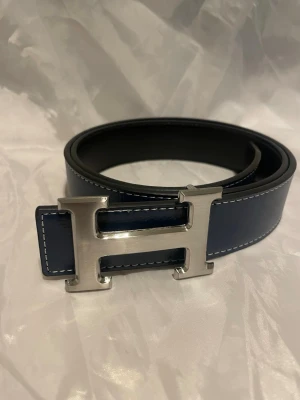 Blått läderbälte med H-spänne - Snyggt blått bälte i läder med kontrastsömmar och ett stort, silverfärgat H-spänne i metall. 110cm. Pris går att diskutera 