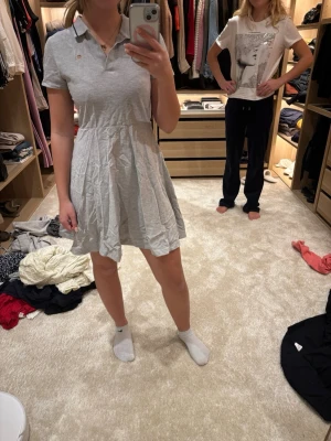 Ljusgrå kort pikéklänning - Säljer en ljusgrå kort klänning i pikéstil med krage och knappar framtill. Klänningen har korta ärmar och en utställd kjol som ger en avslappnad och sophi vibe. Perfekt för dig som gillar enkel och sportig stil.