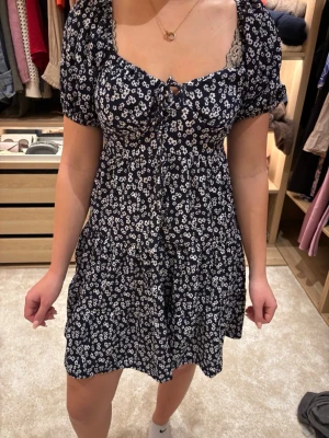 Blommig kortklänning med puffärm - Supersöt kort klänning i mörkblått med vitt blommönster och puffärmar. Klänningen har snörning framtill vid bysten och kan bäras både med puffärmarna uppe eller offshoulder. Tillverkad i mjukt tyg, perfekt för sommarens alla dagar. Ny med prislapp orginalt 399