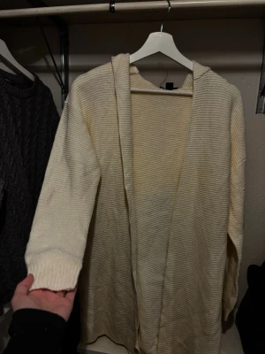 Krämvit stickad hoodiekofta - Mysig krämvit stickad kofta med huva och långa ärmar. Koftan är öppen framtill och har en avslappnad passform, perfekt för lager på lager. Tillverkad i mjukt stickat material som känns skönt mot huden.