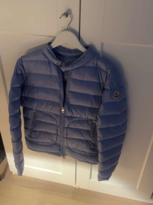 Blå dunjacka från Moncler - Snygg blå dunjacka från Moncler med quiltad design och rund krage. Jackan har dragkedja framtill, två fickor med dragkedja och en liten Moncler-logga på ärmen. Perfekt för kyliga dagar och ger en clean look.