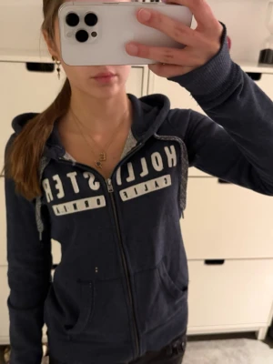 Mörkblå zip up hoodie från Hollister - Mörkblå hoodie med dragkedja från Hollister. Stor vit logga på bröstet, två fickor fram och justerbar huva med grå snören. Mjuk bomullsblandning och ribbade muddar vid ärmslut och nederkant. Perfekt för en avslappnad stil. 2 små defekter visas på sista bilderna!
