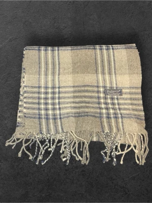 Grå-rutig halsduk från Burberry - Klassisk halsduk från Burberry i beige och blått rutmönster. Tillverkad i mjuk ull med fransar i båda ändar och diskret Burberrys-etikett. Perfekt accessoar för att lyfta din outfit med en tidlös touch.