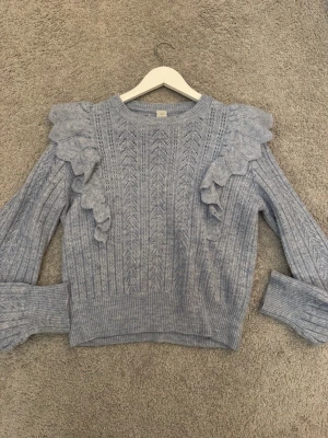 Ljusblå stickad tröja med volang - Supersöt ljusblå stickad tröja med volangdetaljer på axlarna och ett fint hålmönster över hela tröjan. Tröjan har långa ärmar och breda muddar. Perfekt för dig som vill ha något gulligt och trendigt till hösten eller vintern.