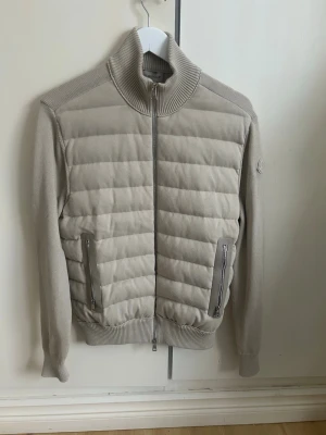 Moncler cardigan - Säljer en beige quiltad jacka från Moncler med ribbad hög krage och muddar. Endast testad, inga skador eller tecken på användning!