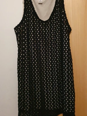 Klänning - Svart virkad kortklänning från Motherhood med ärmlös design och rund hals. Klänningen har ett genombrutet mönster och medföljer ett vitt knytband i midjan för extra detalj.