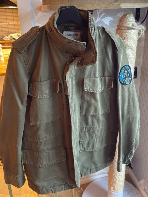 Olivgrön parkajacka Zadig & Voltaire - Säljer en olivgrön parkajacka från Zadig & Voltaire med fyra stora fickor framtill och cool patch på ärmen. Jackan har hög krage, axelklaffar och stängs med knappar. Materialet är bomull och jackan har en avslappnad passform.