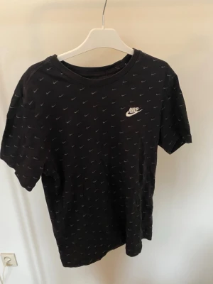 Svart Nike t-shirt med mini-swoosh, Storlek Small - Svart t-shirt från Nike med små gråa swoosh-loggor över hela tröjan och en större Nike-logga på bröstet. Klassisk rund hals och korta ärmar. Tillverkad i 100% bomull för skön känsla och enkel stil.