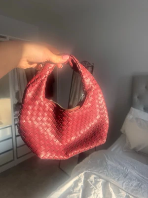 Vinröd flätad axelväska Gina Tricot - Säljer en vinröd axelväska från Gina Tricot i snygg flätad design. Väskan har rundad form, dragkedja upptill och är gjord i syntetmaterial som liknar skinn. Perfekt storlek för det viktigaste och ger en trendig touch till din outfit.