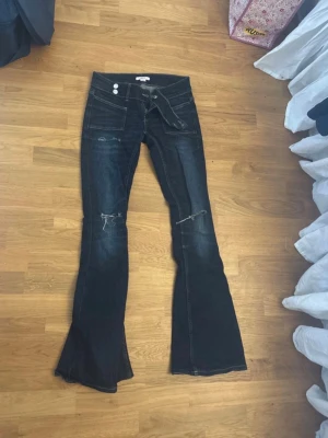 Mörkblå bootcut jeans med slitningar - Säljer ett par mörkblå jeans med bootcut passform och slitna detaljer vid knäna. Byxorna har låg midja, stora fickor fram och dubbla knappar i midjan. Materialet är klassisk jeans och de har en cool, avslappnad vibe.