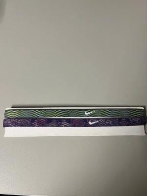 Nike mönstrade hårband 2-pack - Två snygga hårband från Nike med coolt zigzag-mönster. Ett band är i grönt, gult och svart, det andra i lila och blå toner. Båda har vit Nike-logga framtill. Perfekta för att hålla håret på plats under träning eller vardag. Aldrig använda. 
