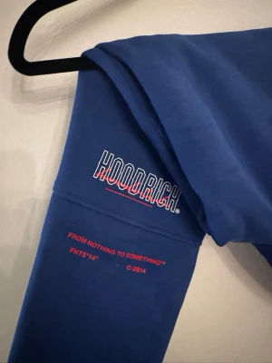 Blå Hoodrich mjukisbyxor med tryck - Säljer ett par blå mjukisbyxor från Hoodrich med stort vitt och rött tryck längs benet och coola detaljer som snörning vid benslut. Knappt använda.