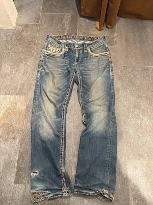 Rock Revival blå jeans Eldon Straight - Säljer ett par Rock Revival Eldon Straight jeans i blå tvätt med slitningar och grova kontrastsömmar. Byxorna har broderade detaljer på fickorna och klassisk femficksmodell. Snyggt slitna kanter och coola broderier bak. Perfekt för dig som gillar statement-jeans.