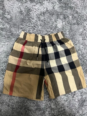 Rutig Burberry shorts med snörning - Säljer ett par klassiska Burberry shorts i beige, svart, vit och röd med det ikoniska rutmönstret. Shortsen har elastisk midja med svart snörning och är tillverkade i ett lätt bomullstyg. Perfekta för sommardagar när du vill ha en clean och stilren look.