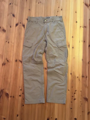 Beiga cargobyxor från Carhartt - Säljer ett par beiga cargobyxor från Carhartt med raka ben och flera praktiska fickor på sidorna och bak. Byxorna har bälteshällor och klassisk Carhartt-logga på bakfickan. Tillverkade i slitstark bomull, perfekta för dig som gillar utility-stil.