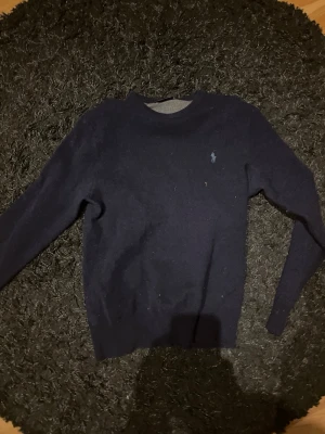 Mörkblå stickad tröja från Polo Ralph Lauren - Klassisk mörkblå stickad tröja från Polo Ralph Lauren med rund halsringning och diskret broderad logga på bröstet. Långärmad modell i mjukt material som passar perfekt till jeans eller chinos.