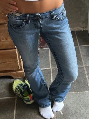 Blå jeans med strassdetaljer - Säljer ett par blå jeans från Y.TWO med snygga strassdetaljer på bakfickorna och coola kontrastsömmar. Modellen har låg midja och bootcut, med klassisk femficksdesign och dekorativa knappar. Perfekta för dig som vill sticka ut lite extra. Skriv till mig för mått!!❤️ jag är 166,5 o de perfekta i löngd för mig endast lite stora i midjan därför det är en vingsak på en sv bilderna men den är bara tillfällig.🥰 