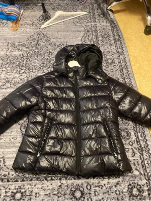 Svart pufferjacka från 157, storlek L - Säljer en svart pufferjacka från 157 i storlek L. Jackan har huva, två snedställda fickor med dragkedja och quiltad design. Materialet är glansigt och känns lätt, perfekt för kyliga dagar. Stängs med dragkedja framtill.