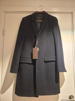 Mörkblå rock från Riley (Brothers) 44 - Säljer en stilren mörkblå rock från Riley (Brothers) med klassisk krage och raka linjer. Rocken har två fickor med lock framtill, två innerfickor och knäppning framtill. Perfekt för dig som vill ha en tidlös och snygg look under kallare månader. Helt ny med prislapp kvar. 