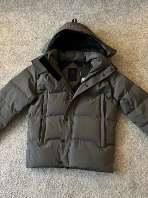 Grå Canada Goose - HELT NY/OANVÄND!!! Säljer en mörkgrå pufferjacka från Canada Goose med huva och diskret logga på ärmen. Jackan är quiltad och har dragkedja samt knappar framtill. Perfekt för kalla dagar och riktigt snygg till streetwear-stil. Först till kvarn!! 