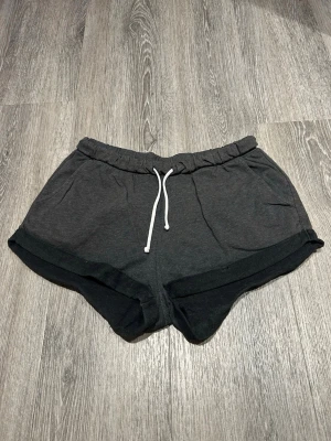 Mörkgrå mjukisshorts med snörning strl. M - Mörkgrå shorts i mjukt bomullsmaterial med vit snörning i midjan. De har en loose passform och uppvikta benslut, perfekta för chill dagar eller träning. Elastisk midja för extra komfort.