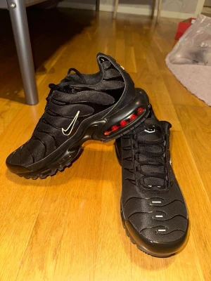 Nike Air Max Plus svarta sneakers - Säljer ett par helt nya svarta Nike Air Max Plus sneakers med vit Swoosh och synlig Air-enhet i sulan med röda och gula detaljer. Ovandelen är i mesh och syntet med vågiga linjer och snörning. Skorna har en chunky sula och sportig vibe, perfekta för dig som gillar streetwear.