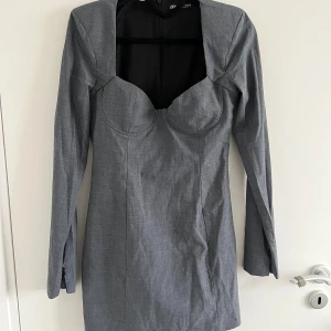 Grå kort klänning från Zara - Snygg grå kort klänning från Zara med markerad byst, lång ärm och fyrkantig urringning. Klänningen har dragkedja bak, stilren look.