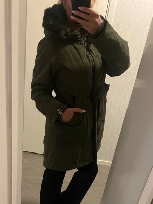 Olivgrön parkas från KappAhl med fuskpäls - Säljer en olivgrön parkas från KappAhl med stor huva fodrad med mjuk fuskpäls. Jackan har dragkedja och knappar framtill, snörning i midjan och två stora fickor. Perfekt för kalla dagar och riktigt snygg till höst- och vinterlooken. Den är supermysig och skön med sin mjuka fuskpäls invändigt i huvan bl a. Riktigt gosig och snygg.