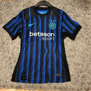 Inter Milan matchtröja Nike Çalhanoğlu - Inter Milan matchtröja med Çalhanoğlu 20 på ryggen med etikett. Tröjan är blå och svart med vertikala ränder, klassisk rund hals och korta ärmar. Nike-logga och klubbmärke på bröstet, tillverkad i lätt och ventilerande Dri-FIT ADV-material. Snygg och sportig design. Skriv vid minsta fundering. Priset kan diskuteras!