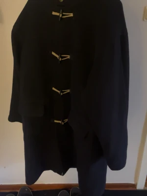 Svart duffel med huva och träknappar - Säljer en svart duffeljacka med huva och klassiska träknappar med snörning framtill. Jackan har lång ärm och en rak, loose passform. Perfekt för dig som gillar tidlös stil och vill ha något som funkar hela hösten och vintern.