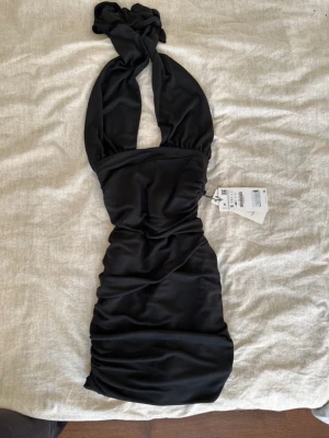 Svart festklänning  - Supersnygg svart kortklänning från Zara med omlott och knyt vid halsen. Klänningen har en tight passform och är tillverkad i ett glansigt polyestertyg som ger en lyxig känsla. Perfekt för dig som vill sticka ut med en trendig look. Dragkedja baktill, helt ny med lappar kvar.