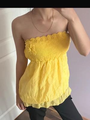 Gul stickad bandeau tubtopp - Supersöt gul bandeau tubtopp med stickad överdel och volangig, luftig nederdel. Toppen har en stor stickad blomma framtill och vågig kant upptill. Perfekt för sommarens alla tillfällen och ger en riktigt glad vibe. Köpt utomlands. Vet inte vilket märke men står lade in italy 