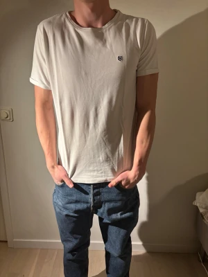 Vit t-shirt med liten logga - Klassisk vit t-shirt med rund hals och korta ärmar. Liten diskret logga på bröstet och enkel, clean design. Tillverkad i mjukt bomullstyg som känns skönt mot huden. Perfekt basplagg för alla tillfällen.