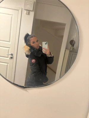 Svart dunjacka med päls från Canada Goose - Säljer en svart lång dunjacka från Canada Goose med ikonisk logga på ärmen. Jackan har huva med fluffig beige päls, knappar längs sidorna och snygga detaljer vid ärmsluten. Perfekt för kalla vinterdagar och riktigt varm.