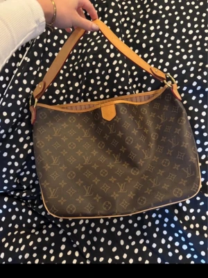  monogram axelväska - Säljer en fin axelväska i brunt monogrammönster med beige och bruna detaljer. Väskan har bred axelrem i skinn med präglad logga, guldfärgade metalldetaljer och rymligt randigt innerfoder med innerficka. Perfekt för dig som vill ha en ikonisk och stilren väska.