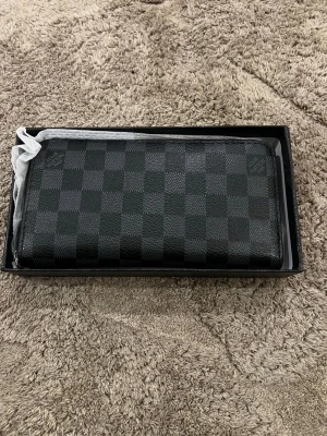  svart rutig plånbok - Snygg svart plånbok med klassiskt rutigt mönster och dragkedja runtom. Tillverkad i skinn med flera fack för kort och sedlar. Perfekt accessoar för dig som gillar stilrena detaljer och exklusiv känsla.