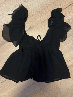 Svart blus med volangärm från H&M - Supersnygg svart blus från H&M med stora volangärmar och knytning framtill. Tillverkad i ett tunt, lätt och lite transparent tyg som ger en luftig känsla. Perfekt för dig som vill ha en trendig och feminin look. Ny utan pris lapp
