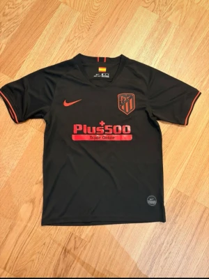 Atlético Madrid Joao Felix Nike tröja - Säljer en svart Atlético Madrid matchtröja från Nike med röda detaljer och Plus500 sponsortryck. På ryggen står det Joao Felix och nummer 7. Tröjan har klubbmärke på bröstet och röda ränder på ärmsluten. Perfekt för fotbollsfans!