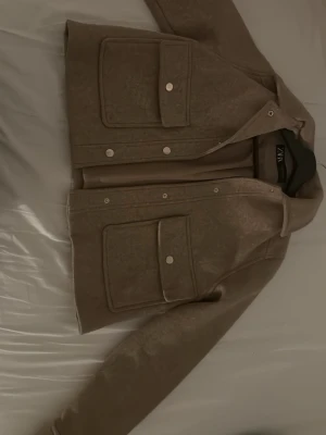 Beige overshirt från ZARA - Säljer en beige overshirt från ZARA med stora fickor framtill och tryckknappar. Jackan har en rak och lite boxig passform, perfekt att slänga över en hoodie eller t-shirt. Materialet känns mjukt och lite tjockare, vilket gör den nice för lager på lager.