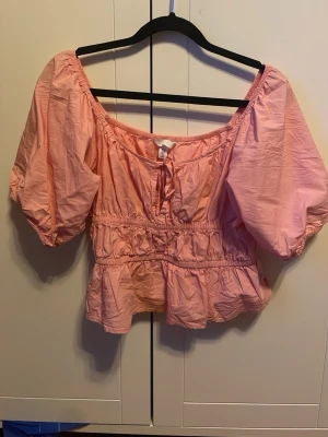 Ljusrosa blus med puffärm - Supersöt ljusrosa blus med puffiga ärmer. Toppen har smock i midjan, volangkant nedtill och en liten rosett framtill. Croppad modell som ger en avslappnad men ändå romantisk vibe. Perfekt till jeans eller kjol! Är i storlek M men passar XS/S