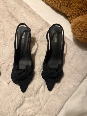 Svarta pumps med rosett  - Säljer ett par eleganta svarta pumps med spetsig tå och låg klack. Skorna har en stor tygrosett framtill och öppen häl med rem. Materialet är troligtvis syntet eller textil, vilket ger en mjuk känsla och stilren look.