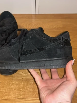 Nike Dunk Low x Undefeated Triple Black - Säljer ett par Nike Dunk Low x Undefeated i helsvart färg. Skorna har ovandel i mocka och mesh, med klassisk Nike swoosh i svart och Undefeated-logga på hälen. Lågt skaft och snörning framtill. Riktigt clean och stilren sneaker för dig som gillar svart på svart.