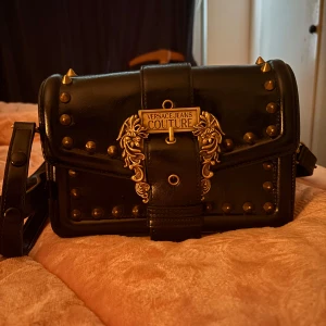 Svart axelväska Versace Jeans Couture - Säljer en svart axelväska från Versace Jeans Couture i skinn med guldiga nitar och detaljer. Väskan har en dekorativ spänne framtill med Versace-logga och coola guldfärgade drakmotiv. Justerbar axelrem och hård, boxig form. Perfekt för dig som gillar statement-accessoarer.