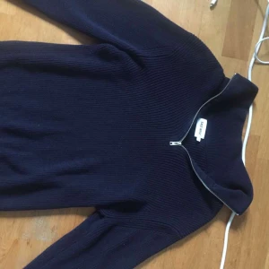 Mörkblå stickad tröja från East West - Mörkblå ribbstickad tröja från East West i storlek M. Tröjan har lång ärm, hög krage och en snygg half zip-dragkedja framtill. Perfekt för dig som gillar en stilren och enkel look med lite edge.