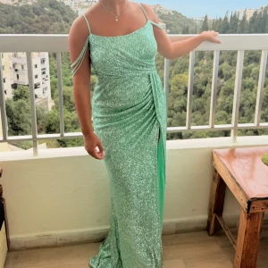 Mintgrön glittrig långklänning - Säljer en mintgrön långklänning med glittrigt tyg och smala axelband. Klänningen har dekorativa pärlband över axlarna, drapering i midjan och en hög slits framtill. Perfekt för bal eller festliga tillfällen när du vill sticka ut. Klänningen är kemtvättad! 