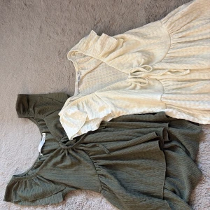 Två toppar från lager 157 - Säljer två toppar från lager 157 i storlek XS. Den ena är vit med knyt i midjan och volangdetaljer, den andra är olivgrön med samma snygga omlott och volang vid ärmarna. Båda har korta ärmar och är gjorda i mjukt, ribbat bomullsmaterial.